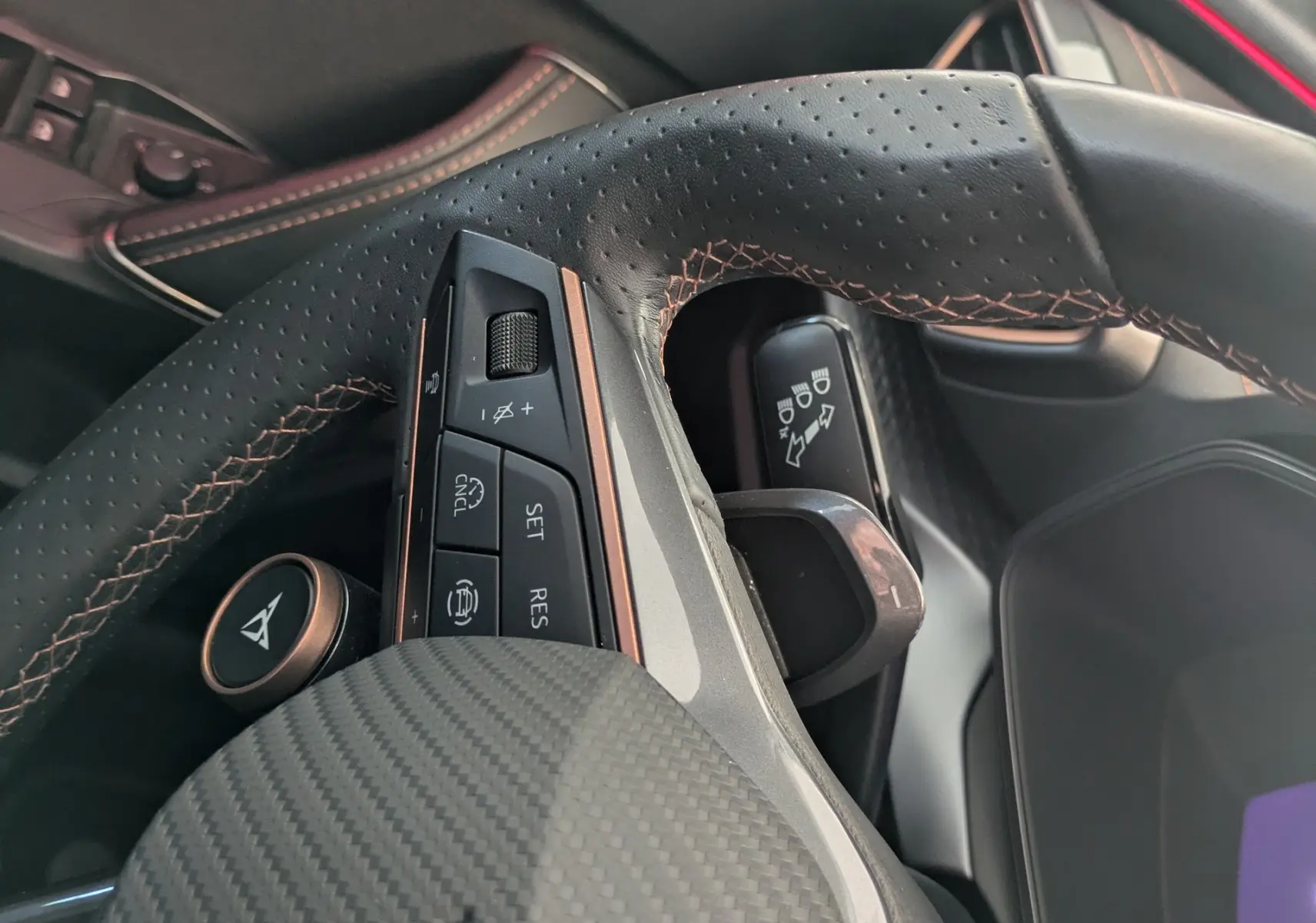 Gros plan sur le volant cuir perforé avec surpiqûres cuivrées et commandes intégrées, intérieur noir avec bande rouge.