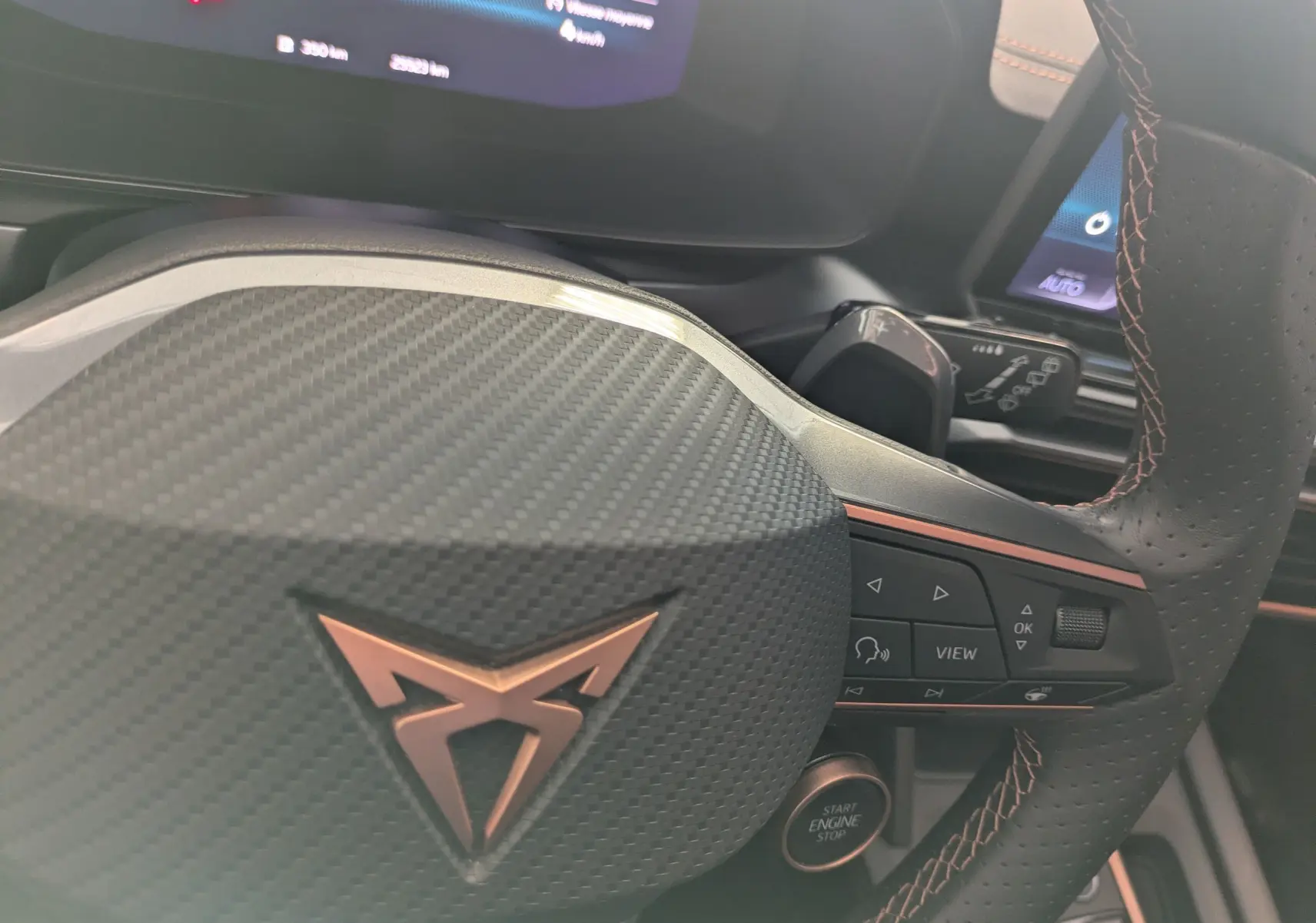 Gros plan sur le volant en cuir perforé avec logo CUPRA cuivré et commandes intégrées, tableau de bord numérique en arrière-plan.