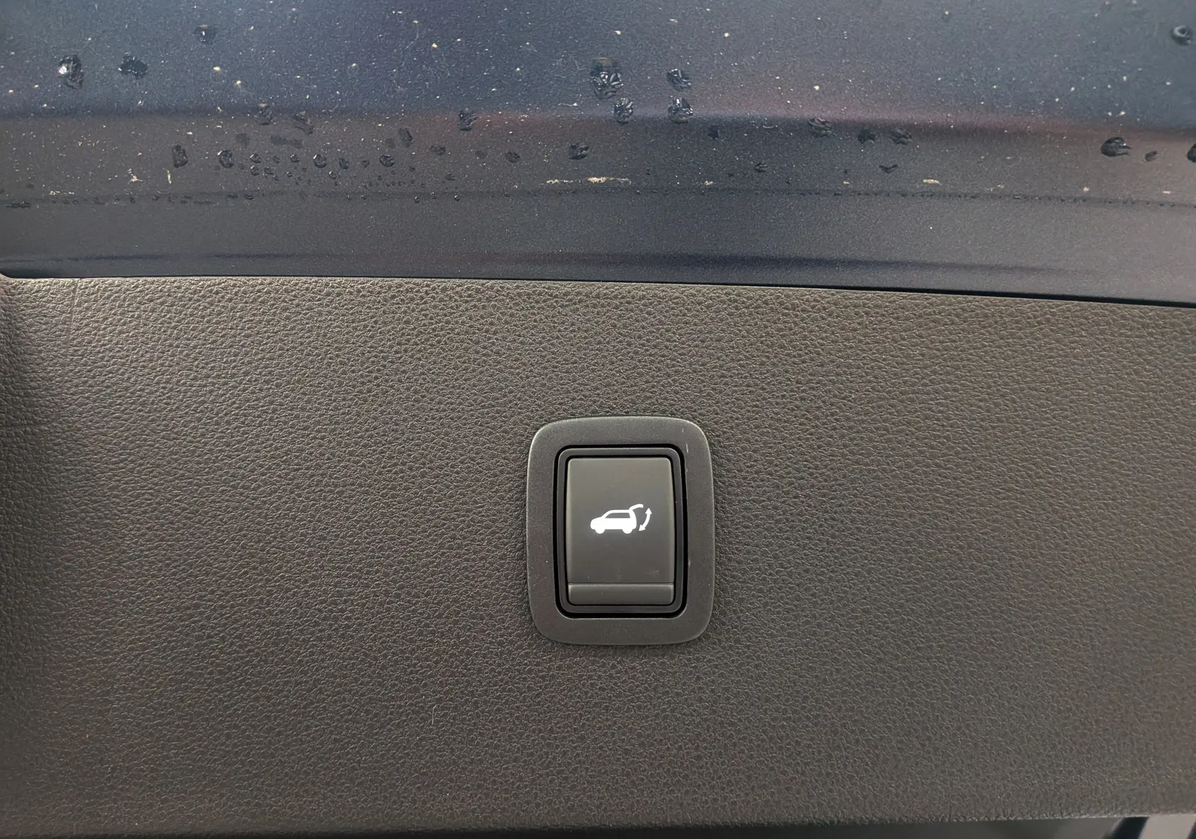 Gros plan sur le bouton d'ouverture électrique du hayon, sur fond intérieur noir du Dacia Bigster Bleu Indigo 2026.