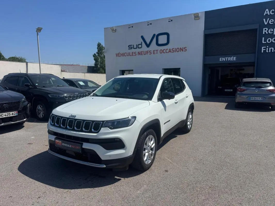 Jeep Compass blanc en 3/4 avant droit, stationné devant un bâtiment de concession sous un ciel bleu clair.