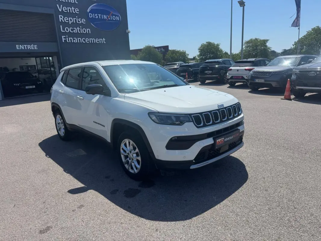 Jeep Compass blanc en vue 3/4 avant droit sur parking extérieur devant un bâtiment commercial.