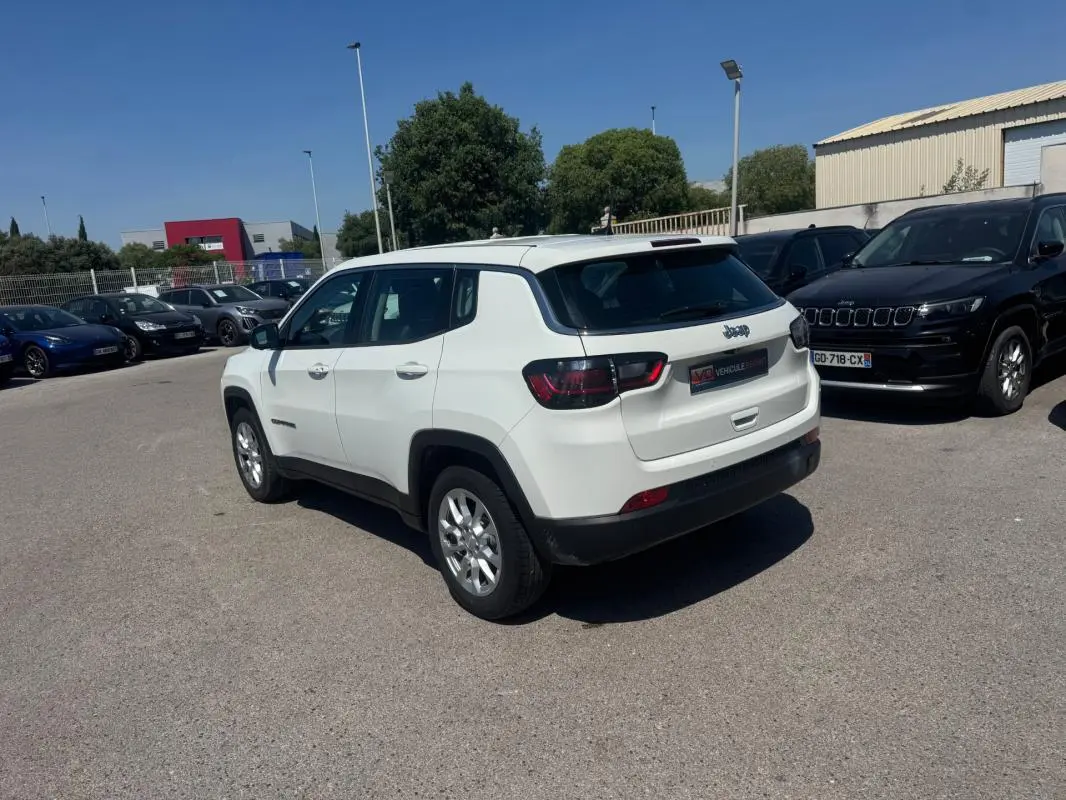 Jeep Compass blanc vu en 3/4 arrière droit sur parking, avec feux arrière sombres et jantes argentées.