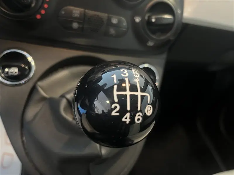 Gros plan sur le levier de vitesses manuel noir brillant de la FIAT 500 blanche, avec marquages des six rapports et marche arrière.