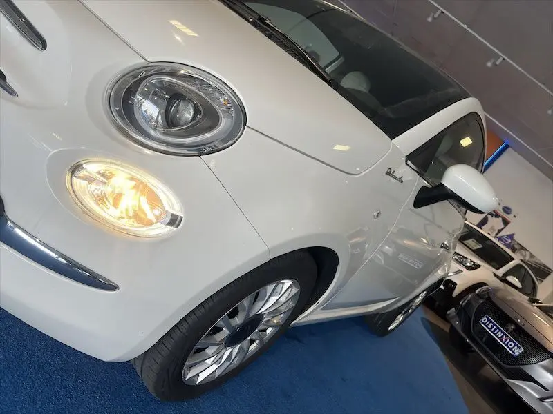 Vue 3/4 avant gauche d'une FIAT 500 blanche avec phares allumés et jantes alliage en showroom.