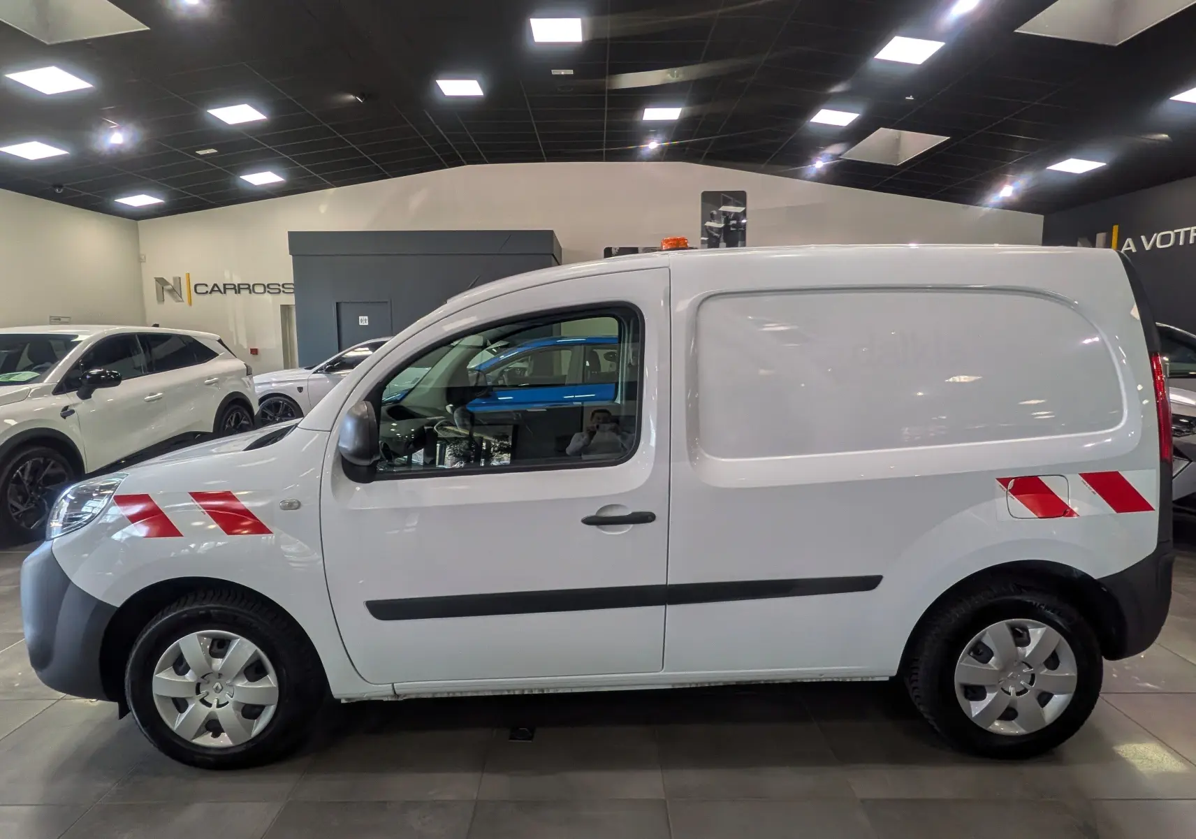 Vue de profil côté gauche d’un Renault Kangoo Express blanc minéral avec bandes réfléchissantes rouges sur ailes avant et arrière.
