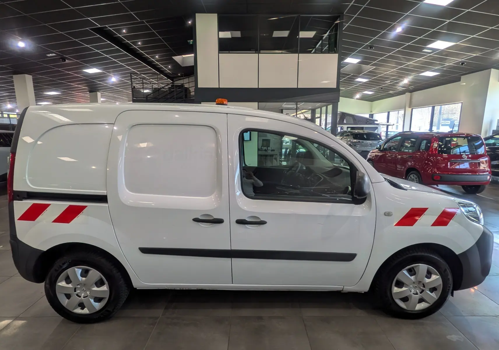 Vue de profil côté droit d’un Renault Kangoo Express blanc minéral avec bandes rouges réfléchissantes et gyrophare orange sur le toit.