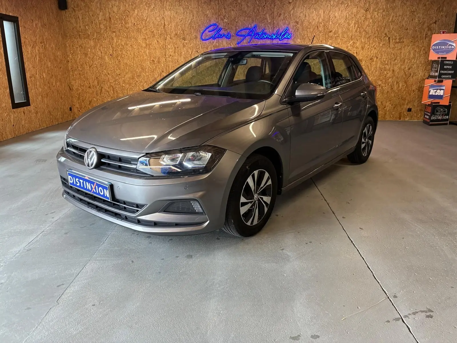 Volkswagen Polo 1.0 TSI gris vue 3/4 avant droit dans un showroom avec éclairage intérieur et logo néon bleu.