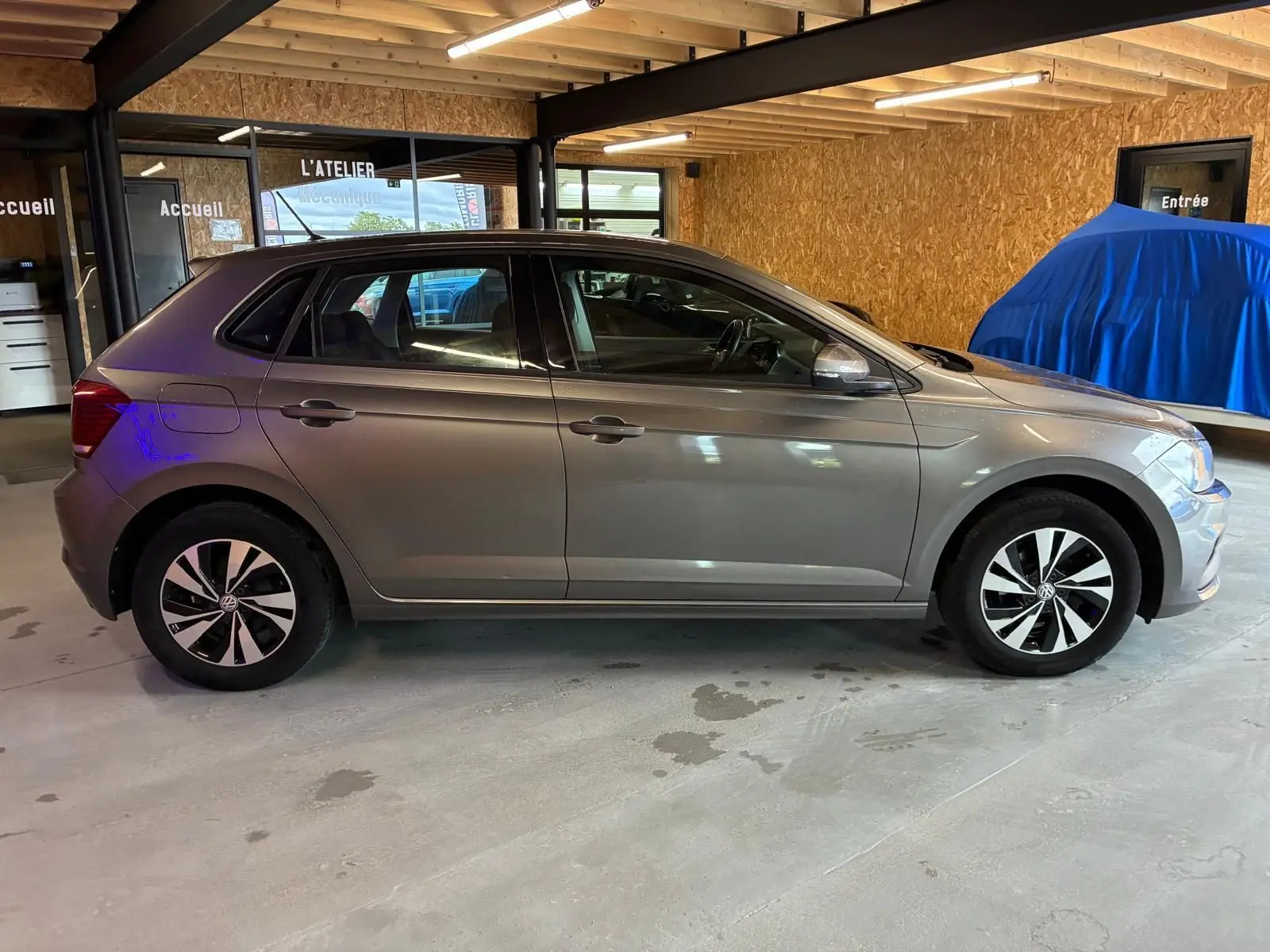 Vue de profil côté gauche d'une Volkswagen Polo gris métallisé 5 portes, stationnée dans un garage avec sol béton.