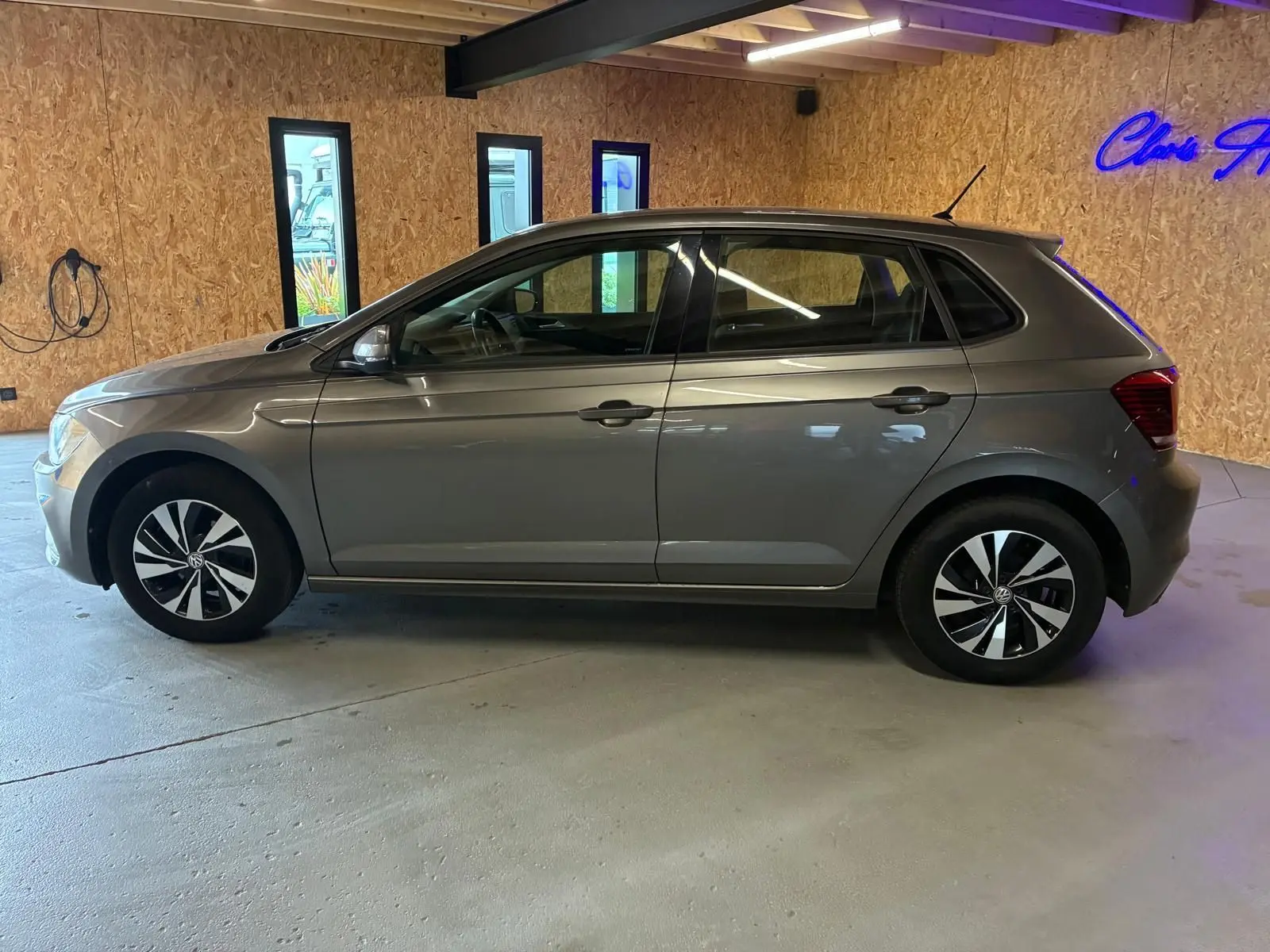 Vue de profil côté gauche d'une Volkswagen Polo gris métallisé 5 portes, avec jantes noires et argentées, en intérieur.