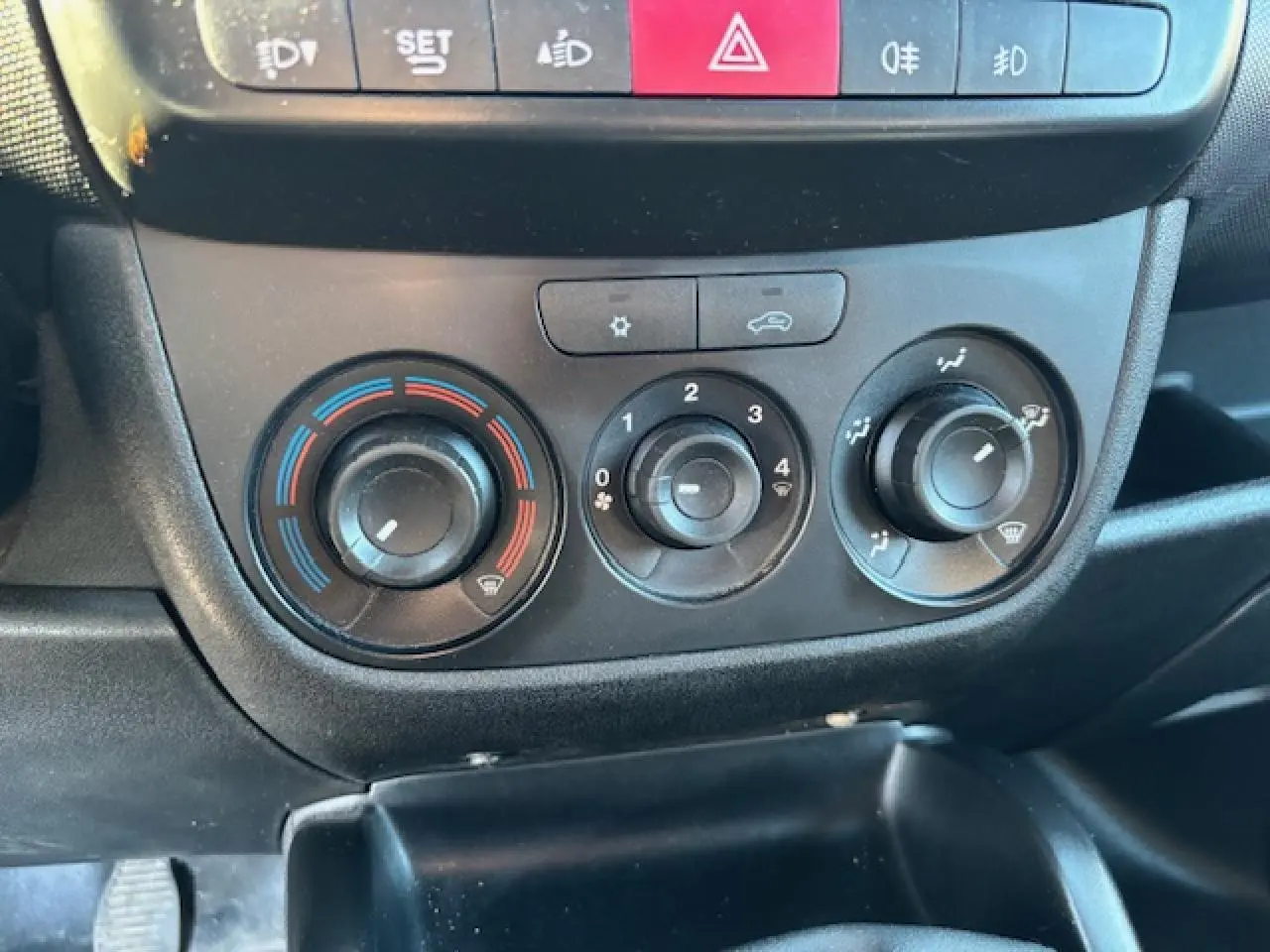 Vue rapprochée des commandes de climatisation manuelle et des boutons de sécurité du tableau de bord d’un Opel Combo blanc de 2015.