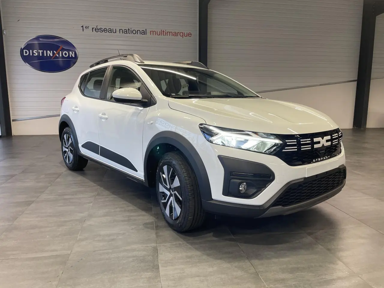 Vue 3/4 avant droit d'une Dacia Sandero Stepway blanche Glacier avec calandre noire et barres de toit grises.