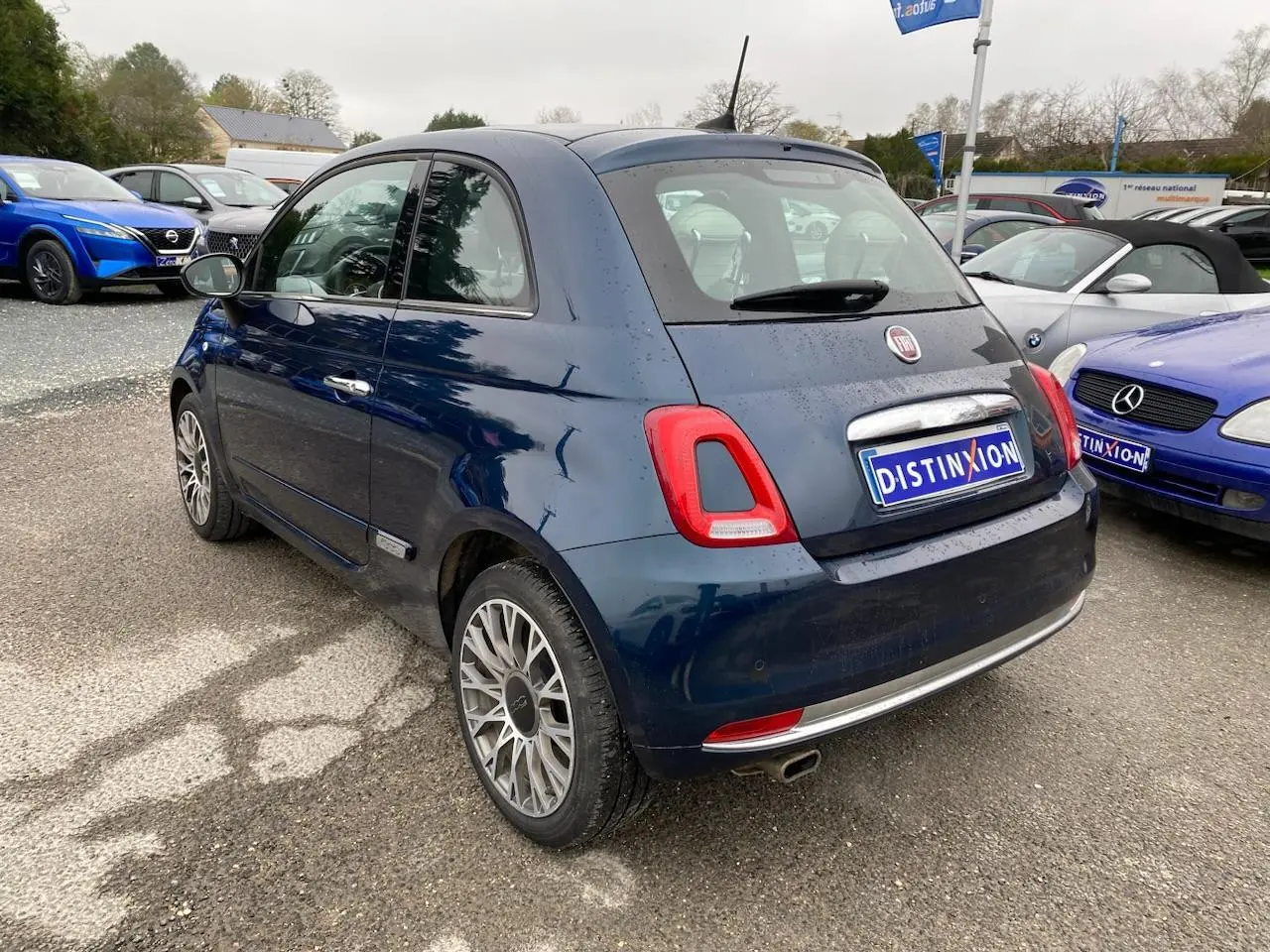 Vue 3/4 arrière droite d'une Fiat 500 bleu foncé avec toit panoramique, stationnée sur un parking extérieur.