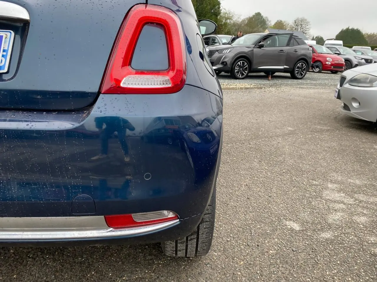 Vue rapprochée arrière gauche d'une FIAT 500 1.2i 69 ECO PACK bleu foncé avec toit panoramique, en extérieur sur parking.