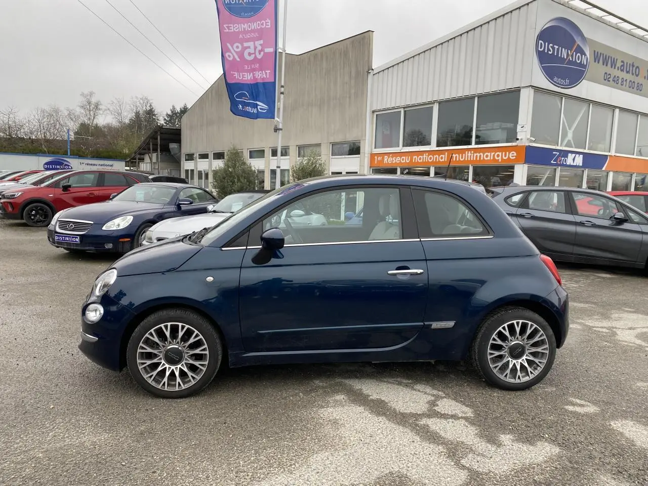 Vue de profil côté gauche d'une Fiat 500 bleu foncé 2019 avec toit panoramique, stationnée devant un concessionnaire.