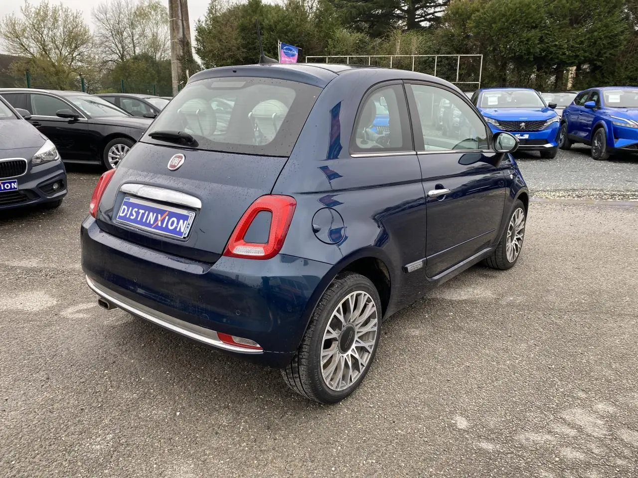 Vue 3/4 arrière droite d'une Fiat 500 bleu foncé avec toit panoramique, garée sur un parking extérieur.