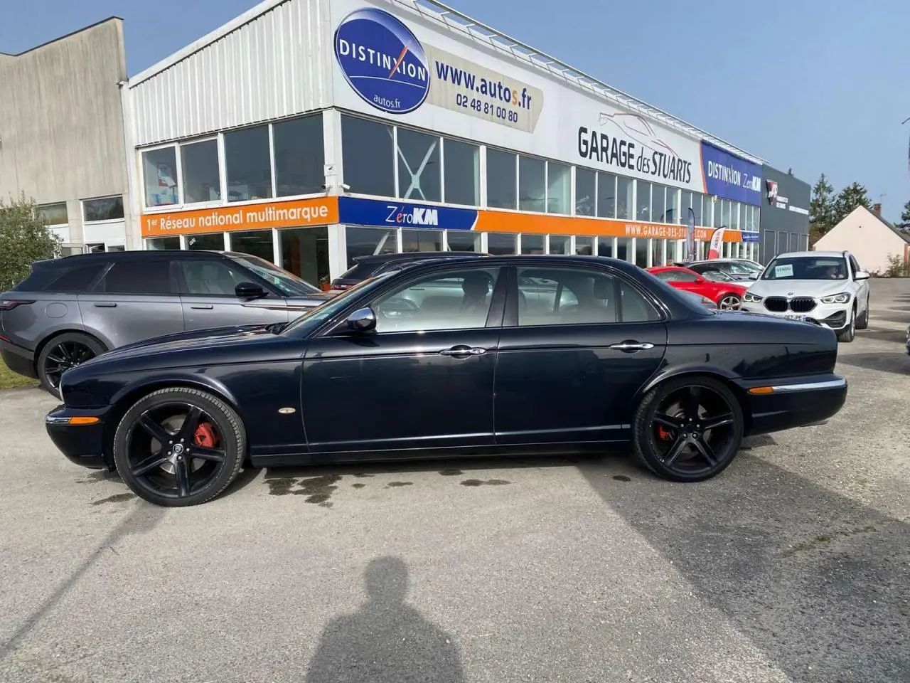Vue latérale gauche d'une Jaguar XJ noire de 2006 avec jantes noires et étriers rouges, garée devant un garage multimarque.