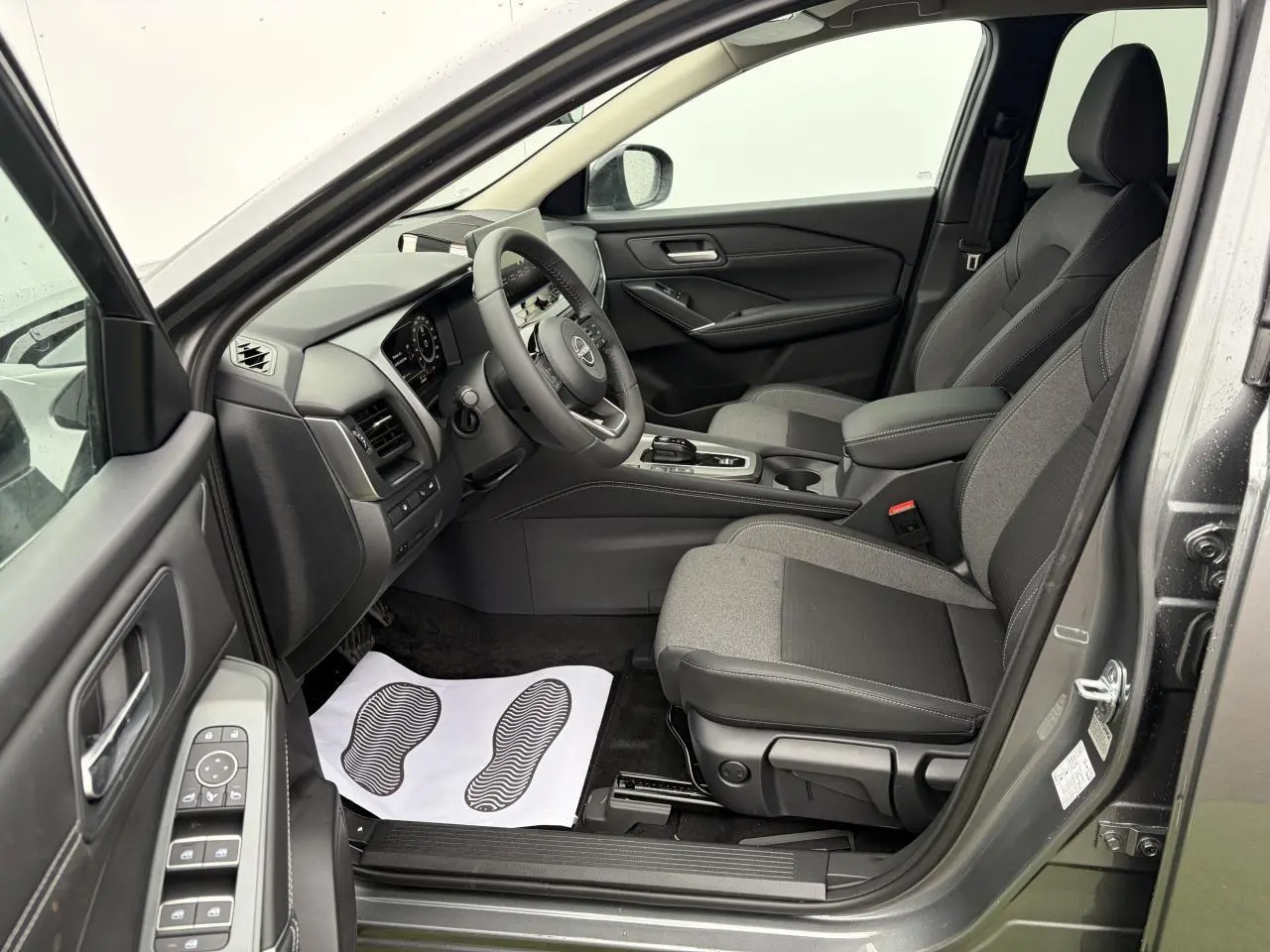Vue intérieure côté conducteur du Nissan Qashqai III 2025 gris foncé, montrant sièges tissu et tableau de bord moderne.