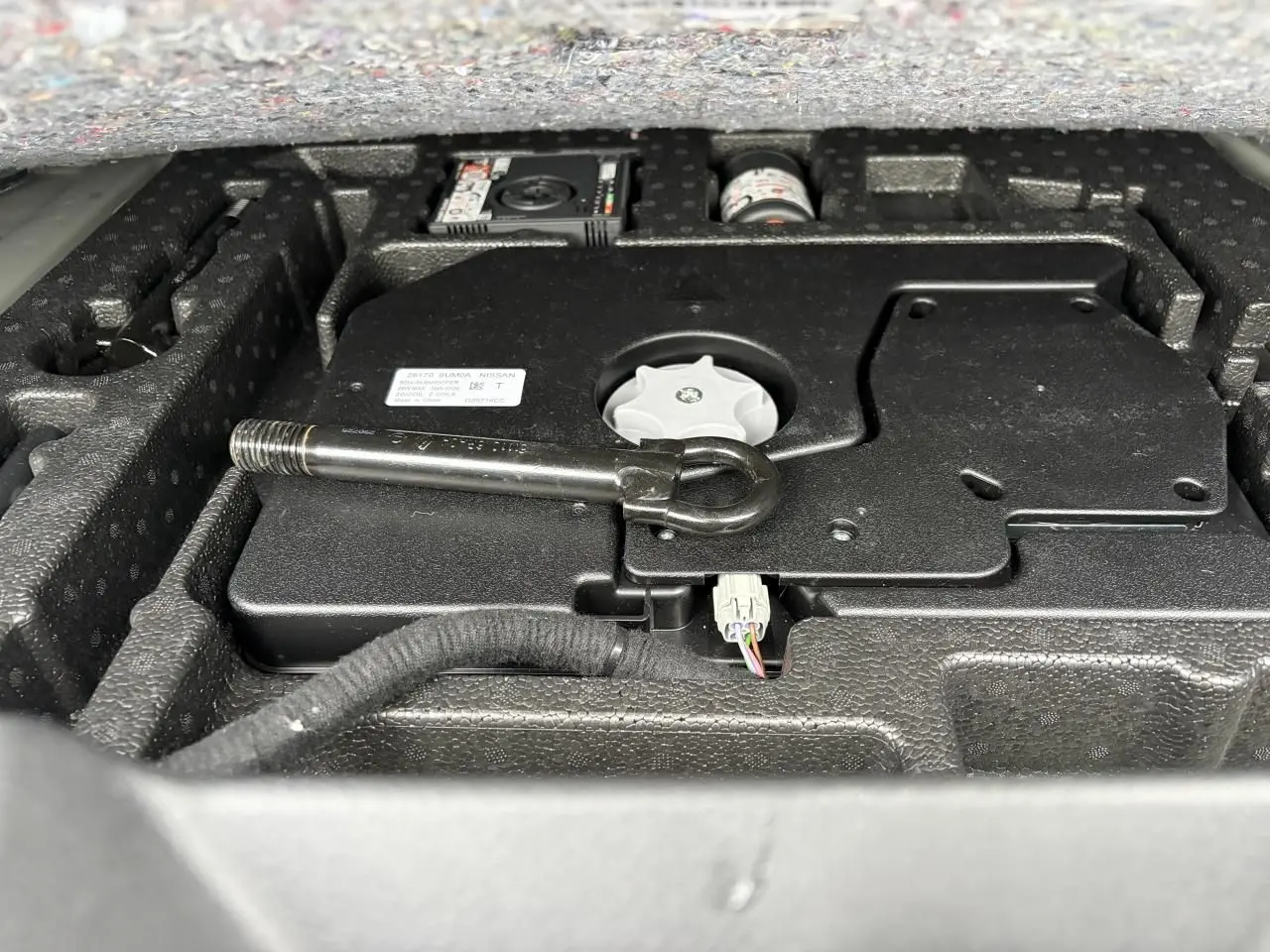 Vue rapprochée sous le plancher du coffre du Nissan Qashqai III gris foncé, montrant l’outillage et le kit de réparation.