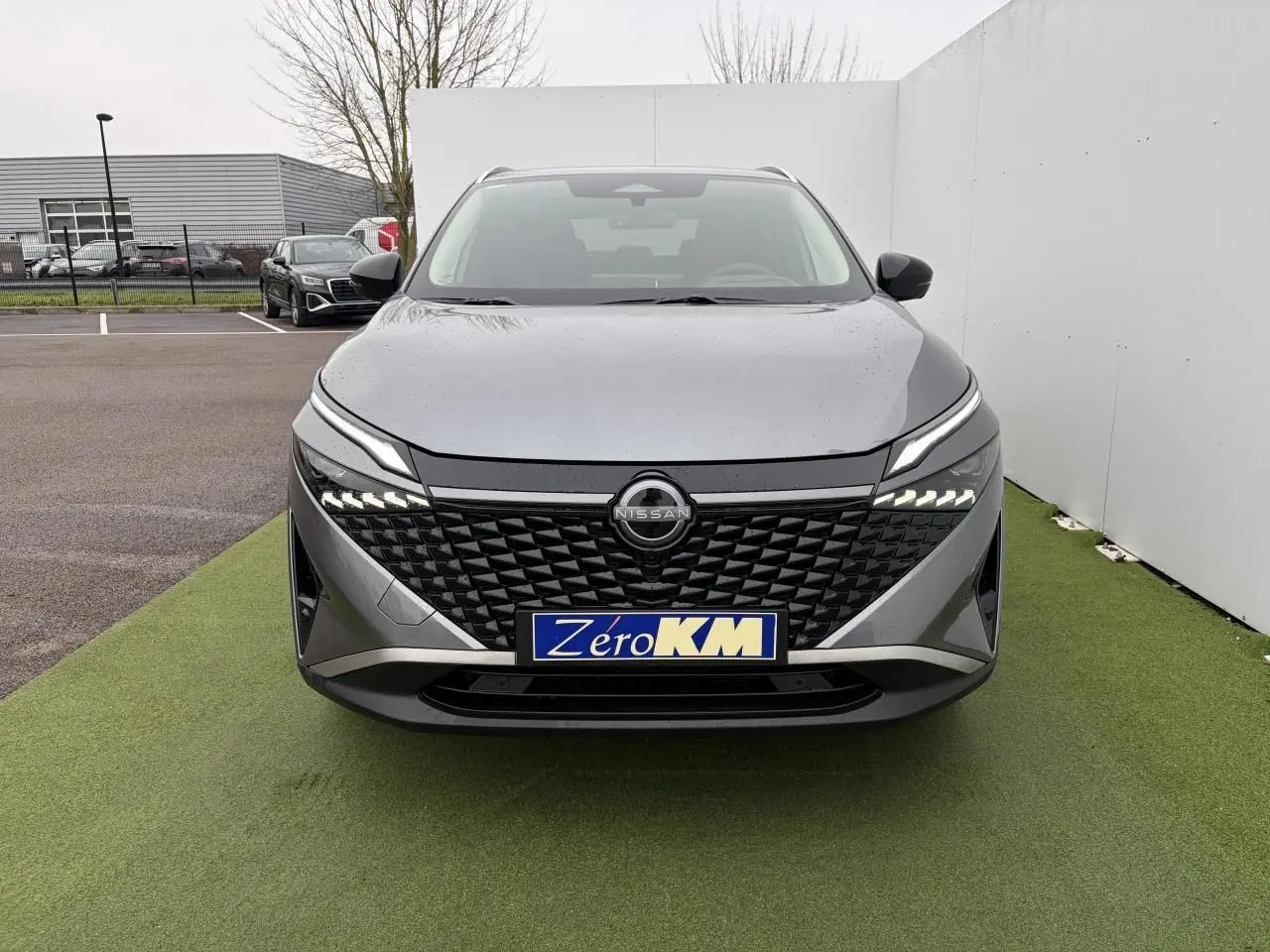 Vue frontale d'un Nissan Qashqai III 1.5 Hybrid E-Power gris foncé métallisé avec calandre noire et phares LED allumés.