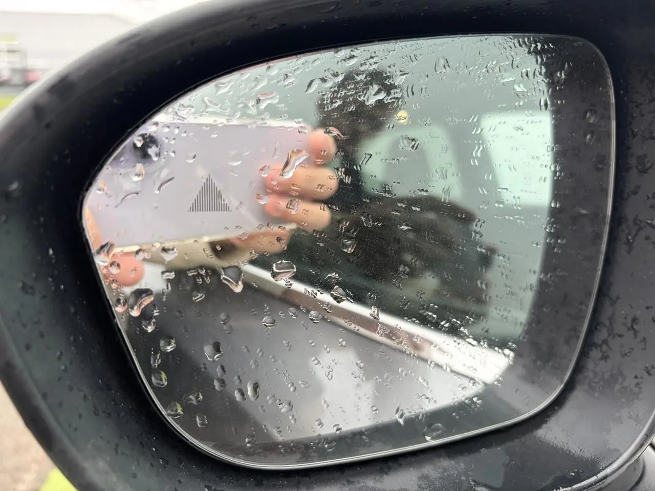 Rétroviseur extérieur droit du Nissan Qashqai gris foncé avec gouttes de pluie et signal d'angle mort visible.