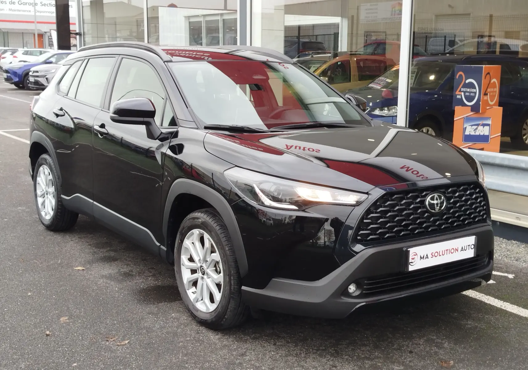 Toyota Corolla Cross noir intense vue 3/4 avant droit, avec calandre noire et feux LED allumés, stationnée devant un showroom.