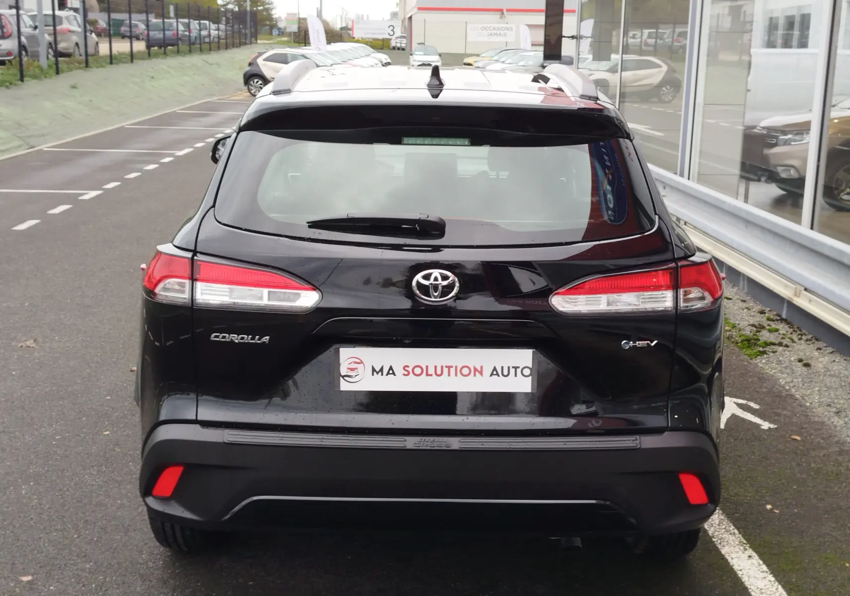Vue arrière d’un Toyota Corolla Cross noir intense, avec feux arrière LED et logo hybride visible sur un parking extérieur.