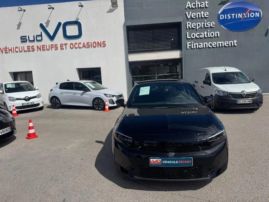 Opel Corsa 1.2 Turbo Hybrid noir vue de face, stationnée devant un garage avec panneaux de vente visibles.