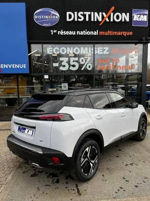 Vue 3/4 arrière droite d'un Peugeot 2008 GT blanc avec toit noir et jantes distinctives, stationné devant un concessionnaire multimarque.