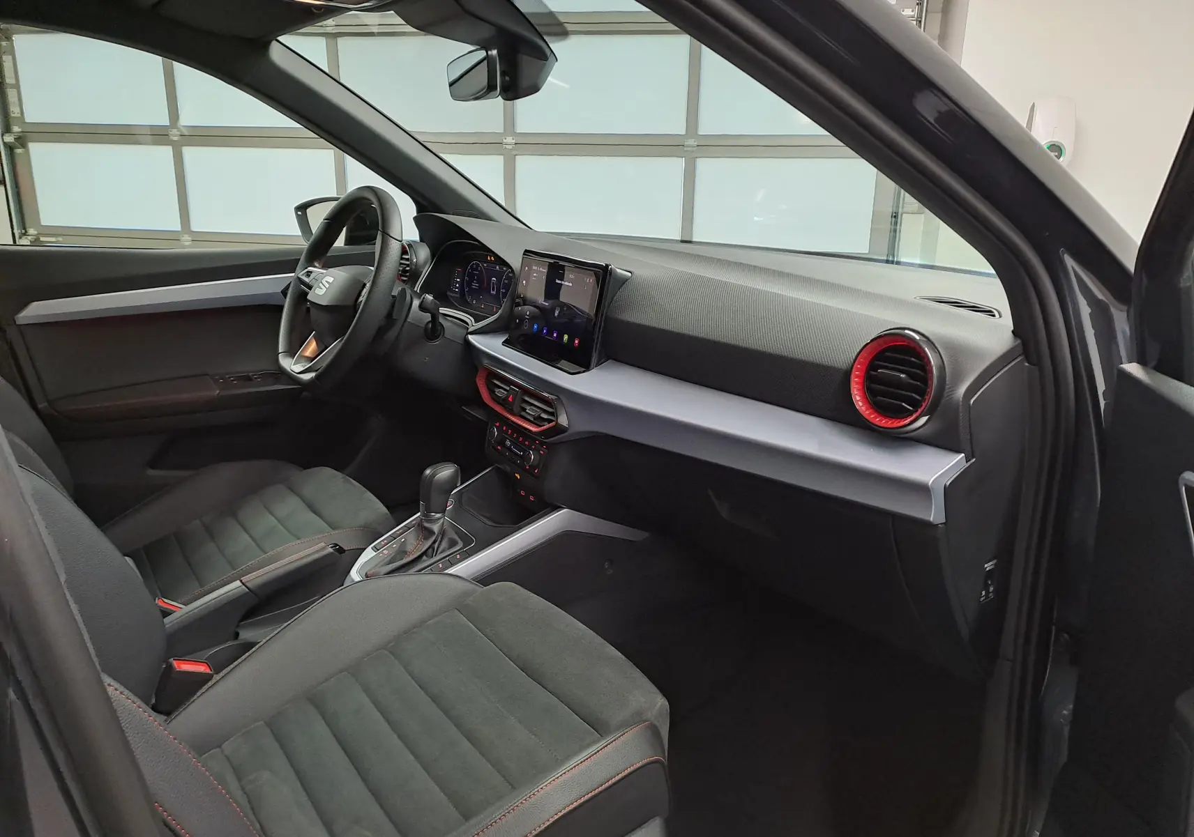 Intérieur de la SEAT Arona 2024 vue côté conducteur, avec sièges noirs, tableau de bord moderne et aérateurs rouges.