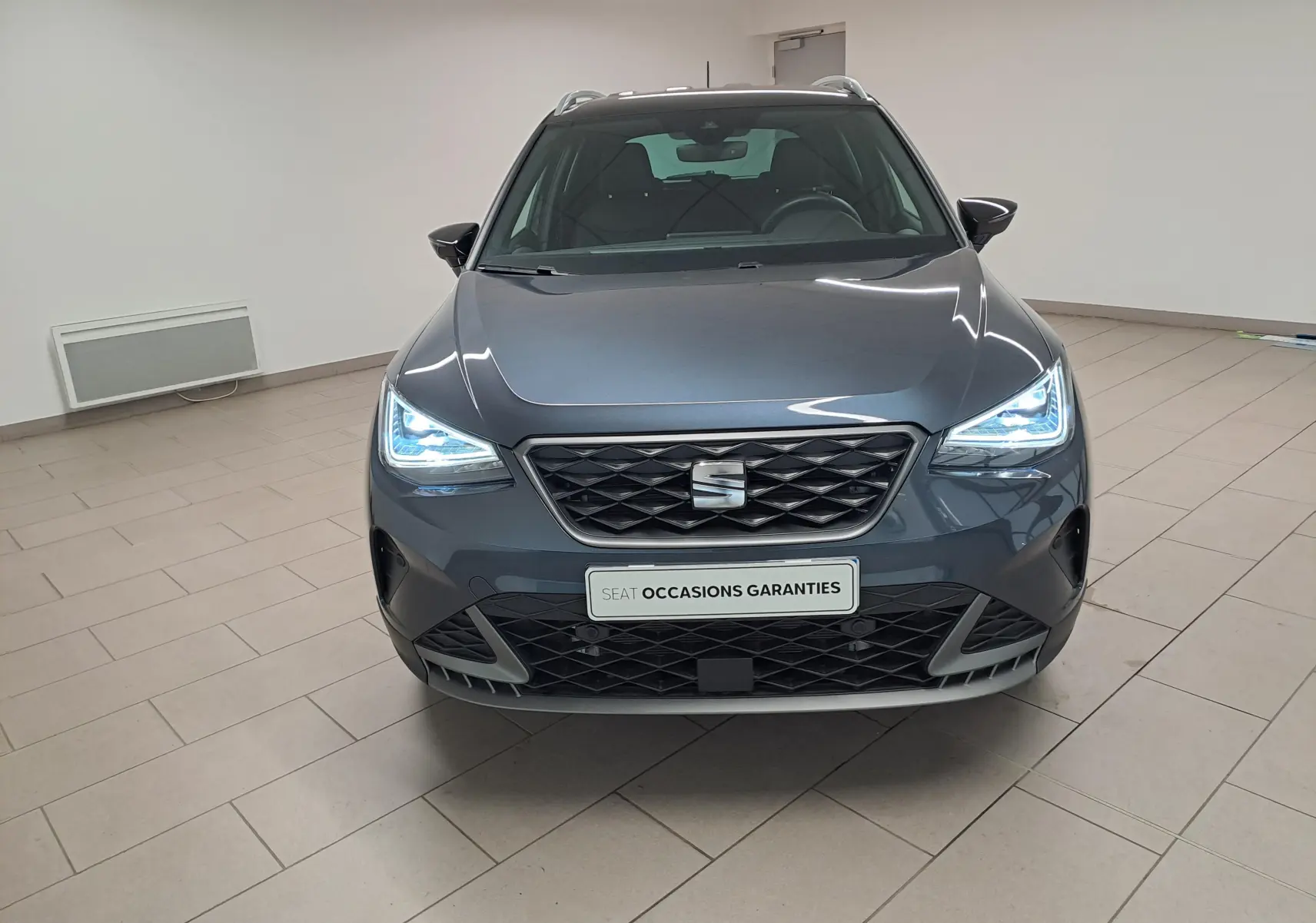 Vue frontale du SEAT Arona 2024 gris magnétique avec toit noir, phares LED allumés dans un showroom.