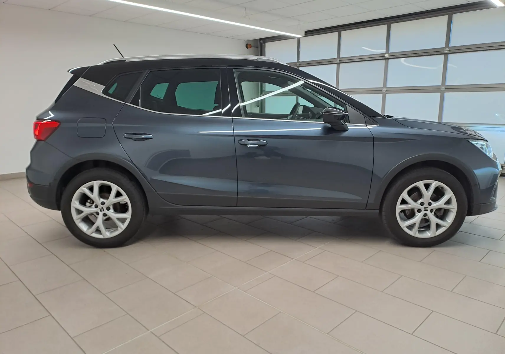 Vue de profil côté gauche d'un SEAT Arona 2024 gris magnétique avec toit noir dans un intérieur lumineux.