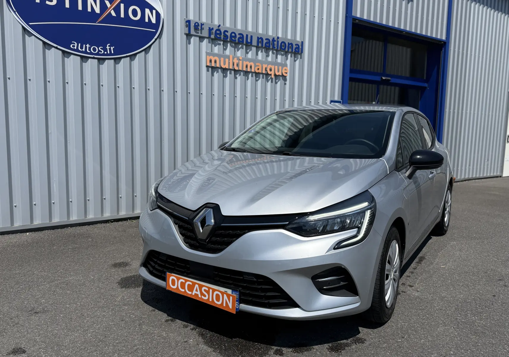 Renault Clio V gris en 3/4 avant droit, avec calandre noire et feux LED, stationnée devant un bâtiment métallique.