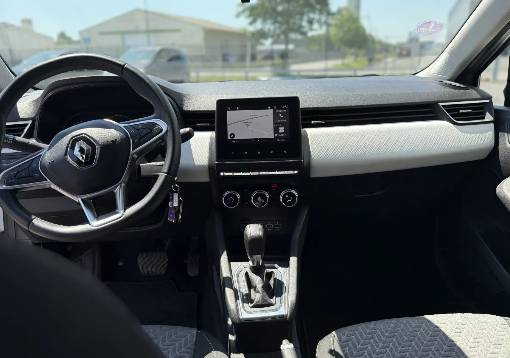 Vue intérieure avant de la Renault Clio V gris, montrant le volant, l’écran tactile central et la console avec boîte automatique.