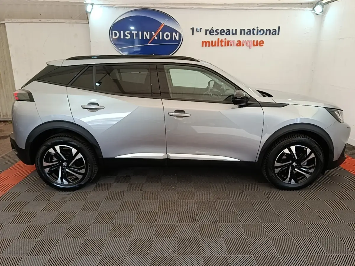 Profil côté gauche d’un Peugeot 2008 gris clair 2022 avec toit noir et jantes alliage bicolores, en intérieur showroom.
