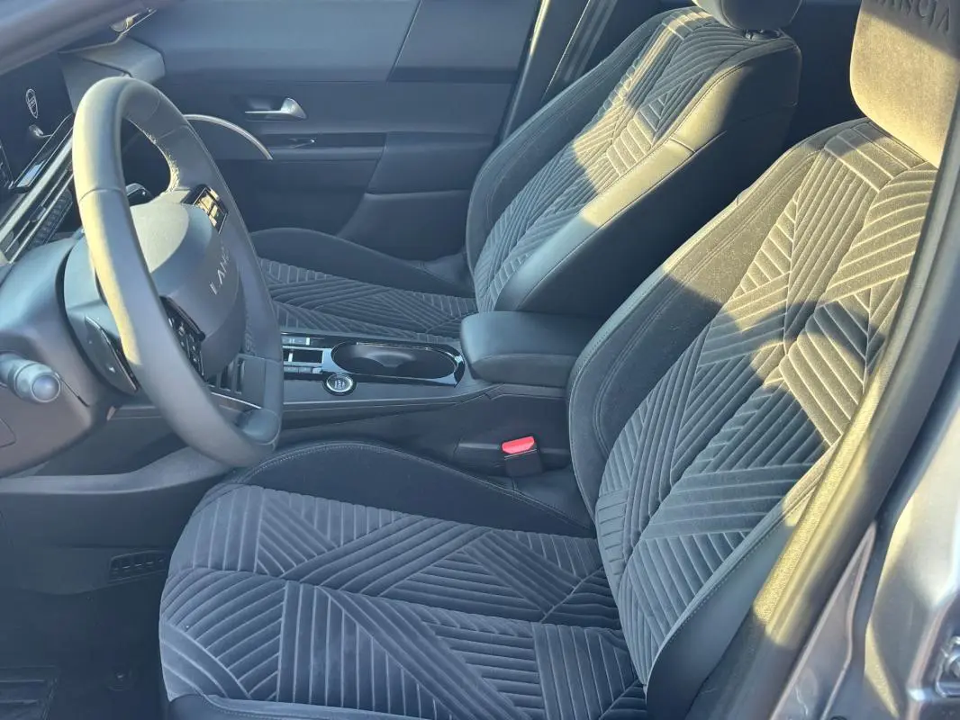 Intérieur avant droit de la Lancia Ypsilon gris 2025, sièges en tissu à motifs géométriques et volant sport visible.