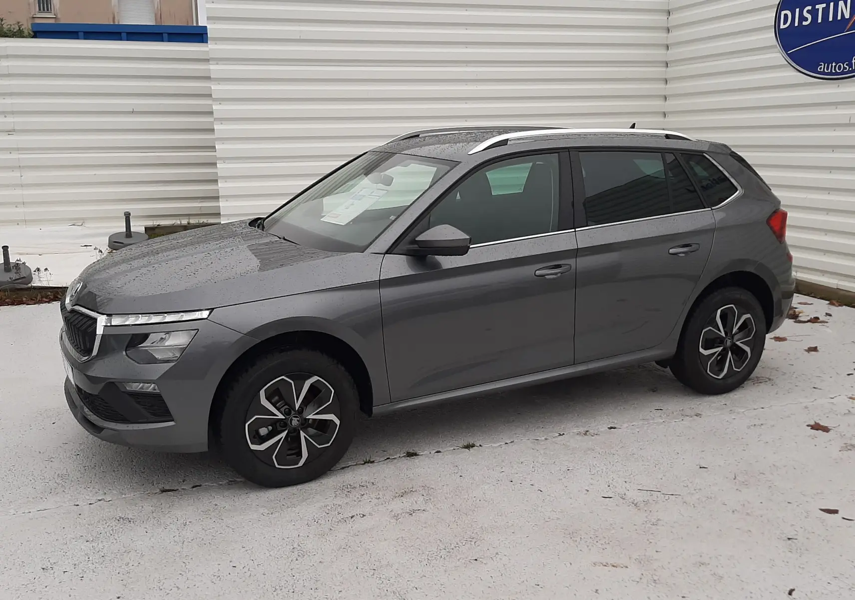 Vue 3/4 avant droit d'un Skoda Kamiq gris graphite métallisé avec jantes alliage et barres de toit argentées.