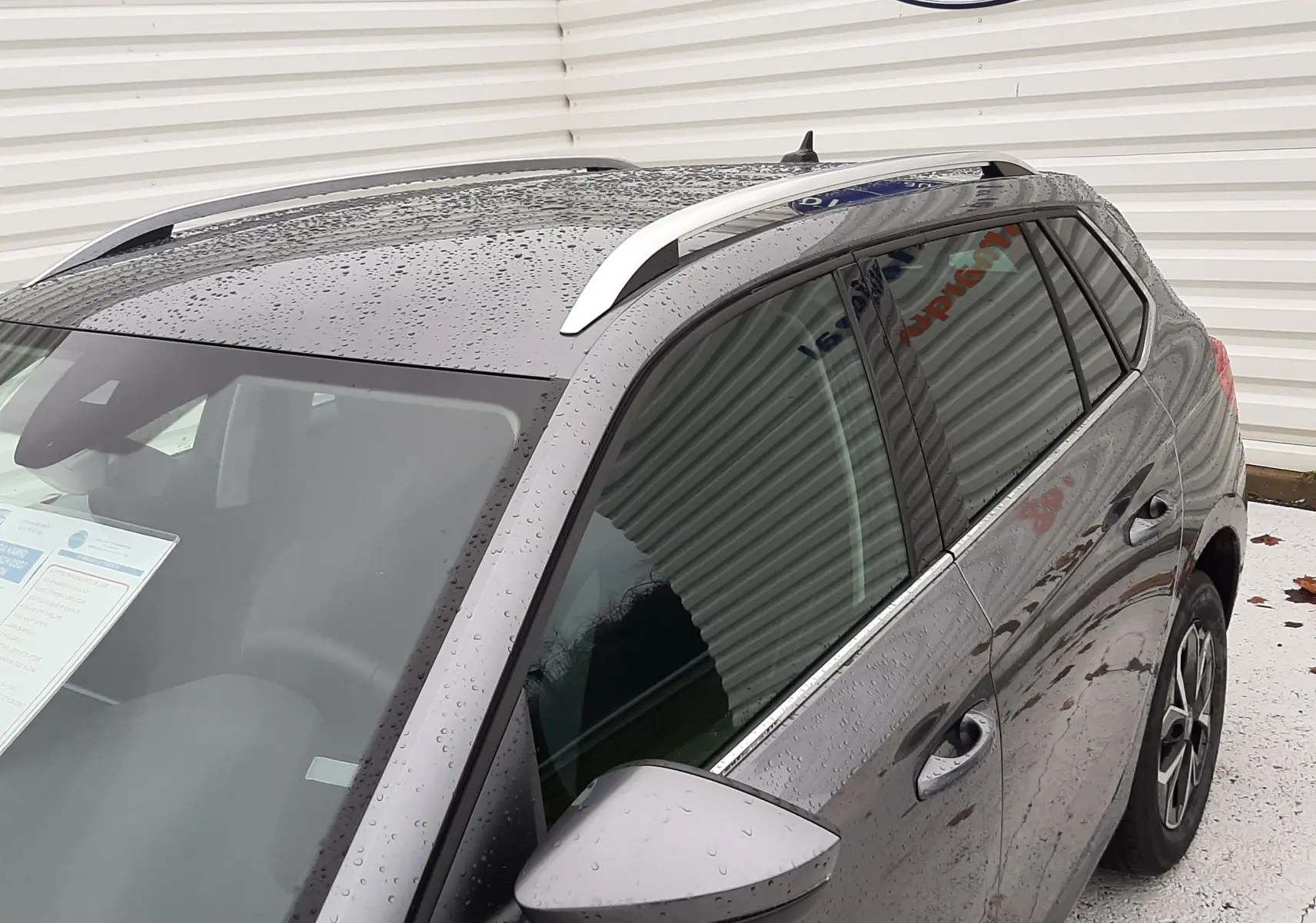Vue de trois quarts arrière droit d'un Skoda Kamiq gris graphite métallisé avec barres de toit argentées sous la pluie.