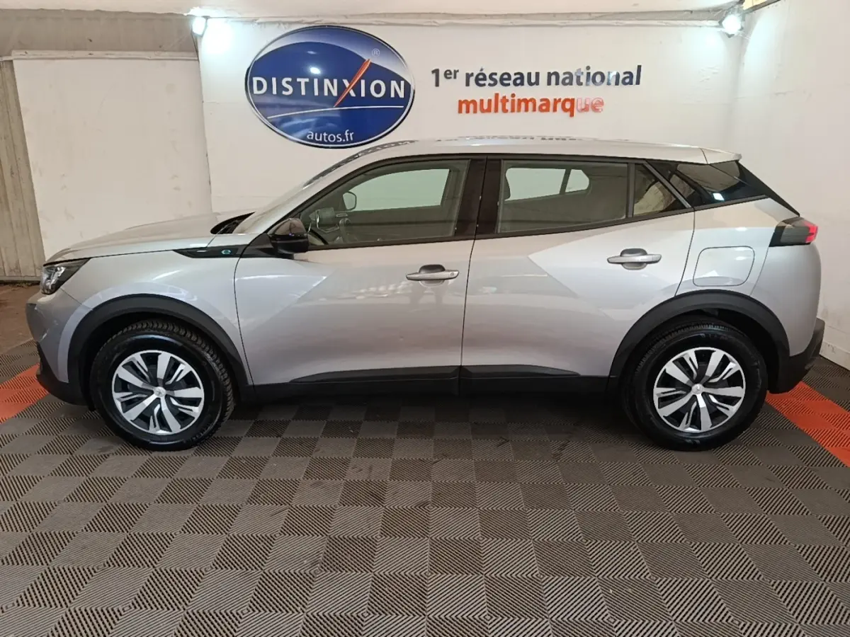 Vue latérale droite d’un Peugeot E-2008 électrique Active Pack gris clair dans un showroom intérieur.