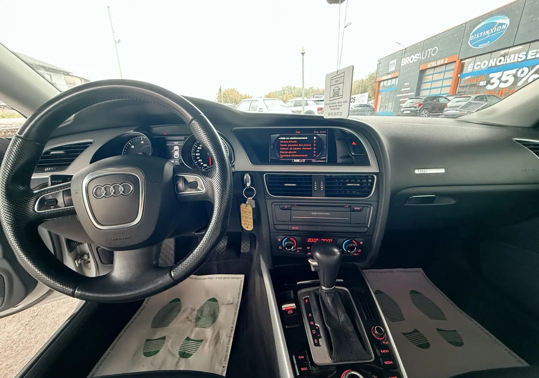 Vue intérieure de l'Audi A5 gris, tableau de bord et volant avec commandes, console centrale et levier de vitesses automatique visibles.