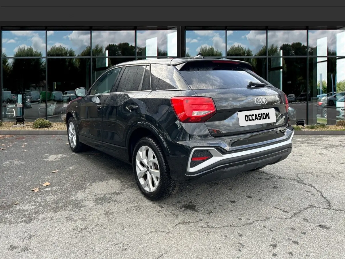 Audi Q2 noir vue 3/4 arrière droit, avec feux arrière rouges et jantes argentées, stationné devant un bâtiment vitré.