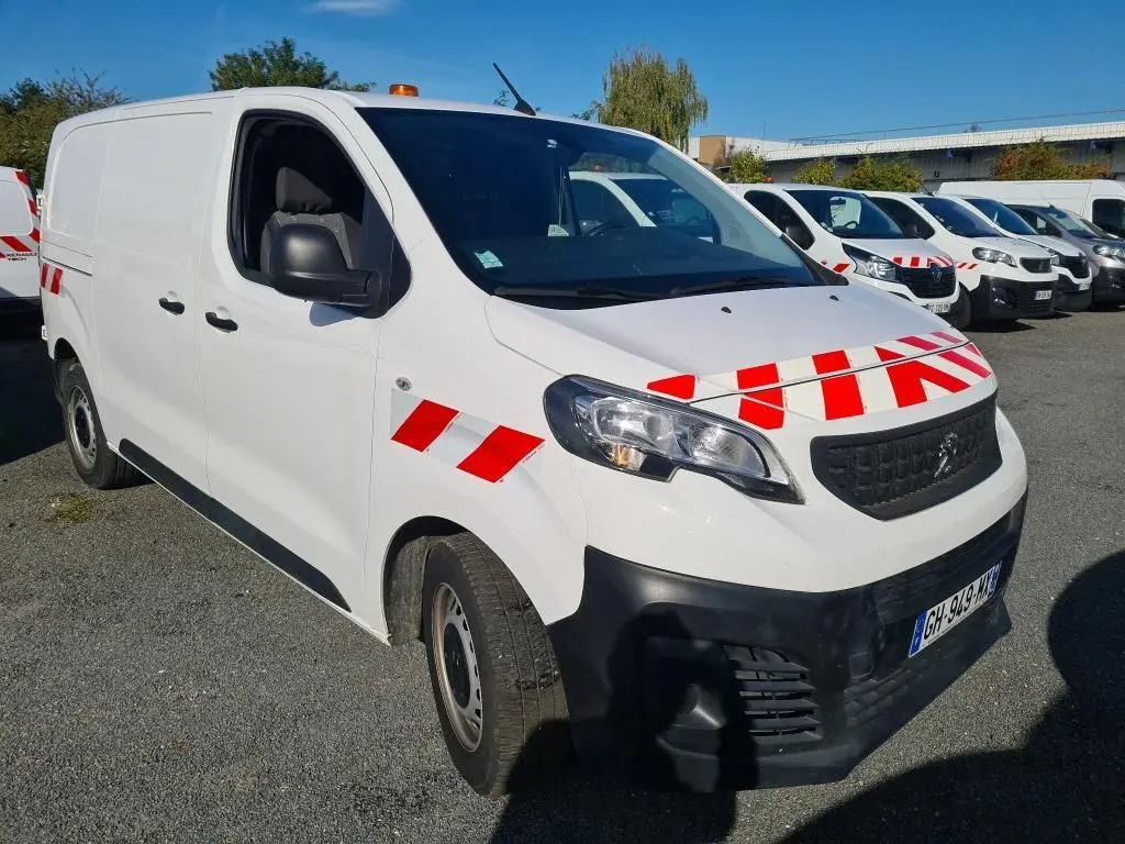 Vue 3/4 avant d'un Peugeot Expert Fourgon blanc avec bandes rouges réfléchissantes et gyrophare orange sur le toit.