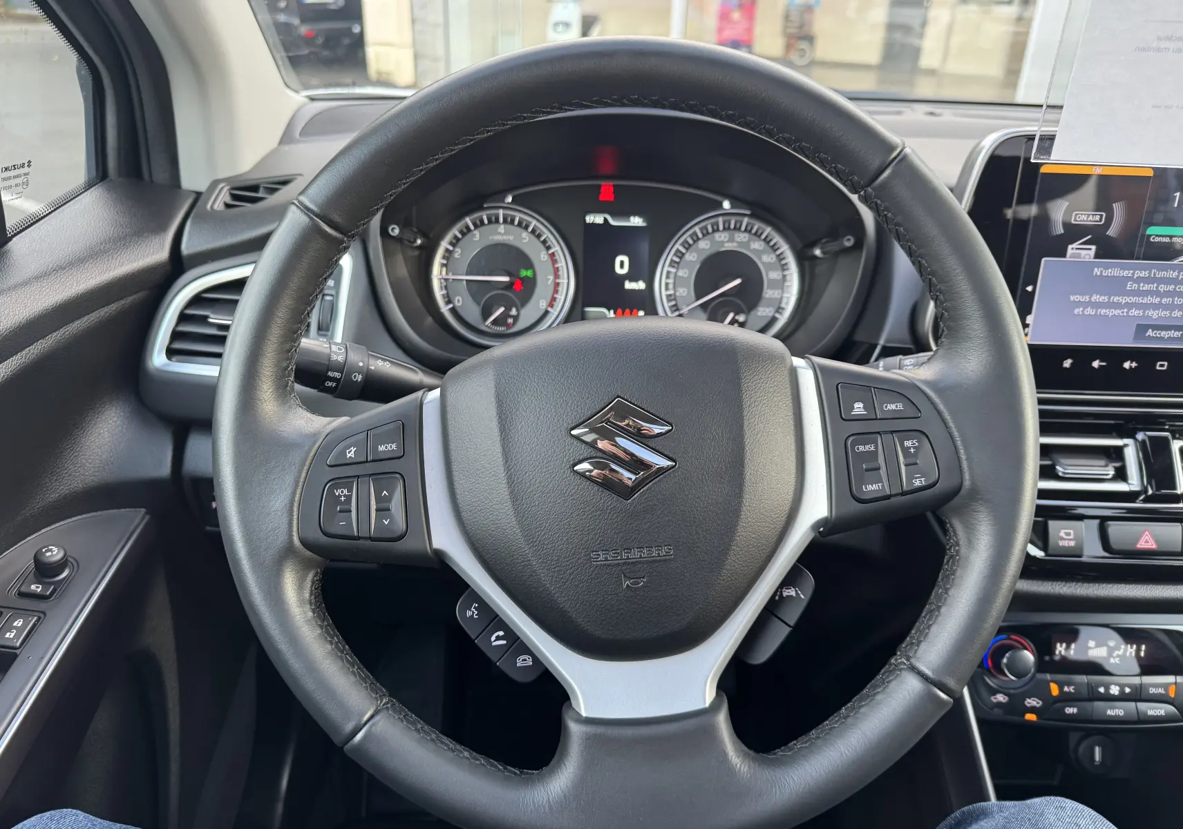 Vue rapprochée du volant noir de la Suzuki S-Cross 1.4 Style Hybrid 2023 avec commandes intégrées et tableau de bord visible.