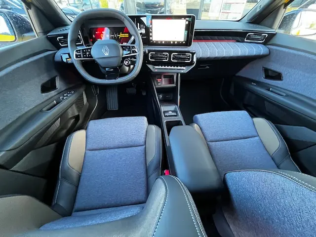 Vue intérieure avant de la Renault R5 Techno 2024, tableau de bord moderne avec écran tactile central et sièges tissu gris.