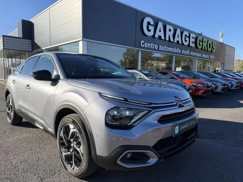 Citroën C4 gris acier vue 3/4 avant droit, avec calandre distinctive et jantes noires, devant un garage multimarques.