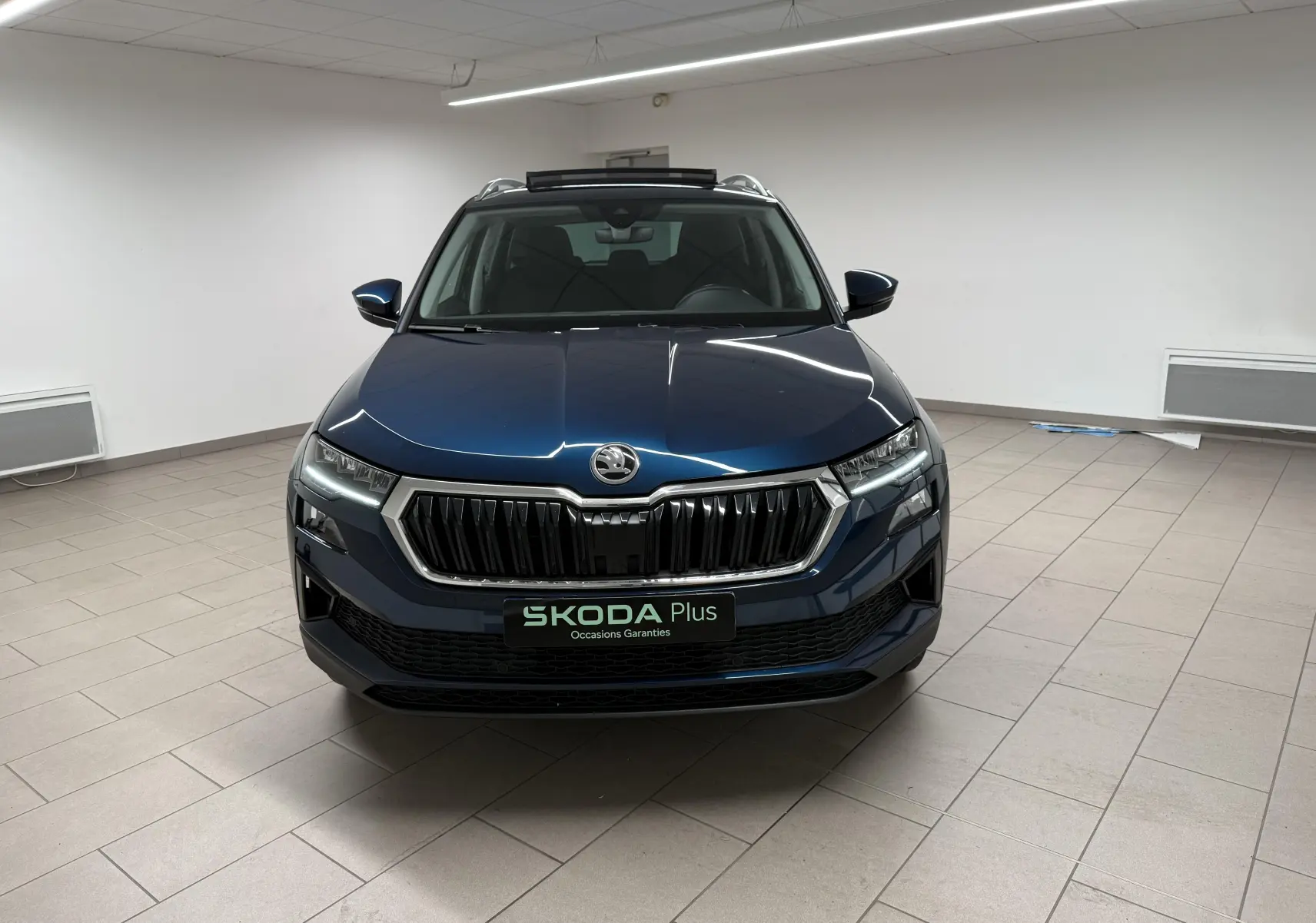 Vue frontale d'un Skoda Karoq 2023 bleu lave avec calandre chromée et toit ouvrant en intérieur showroom.