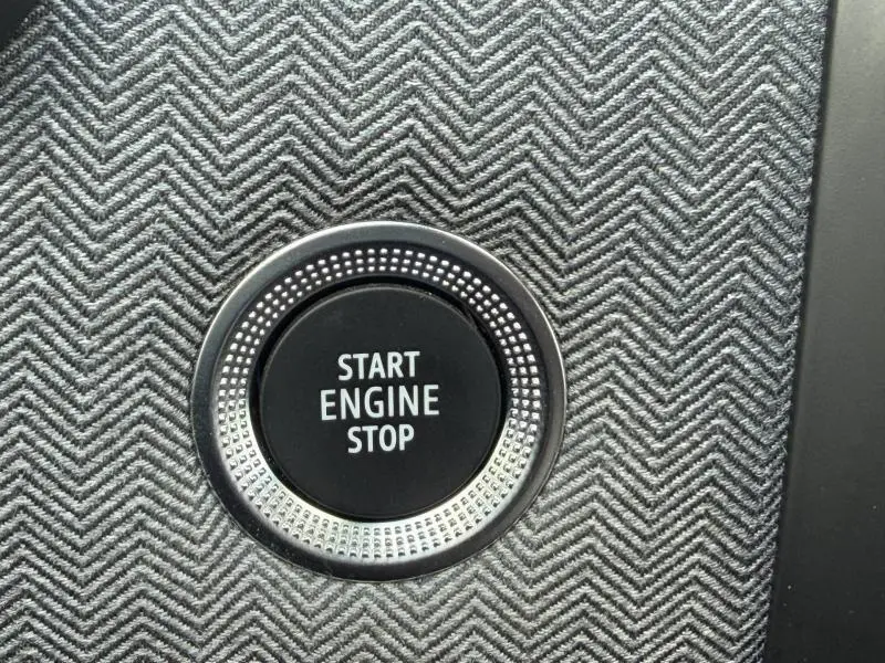 Gros plan sur le bouton Start Engine Stop entouré d’un cerclage métallique, sur un revêtement textile gris à motif chevrons.