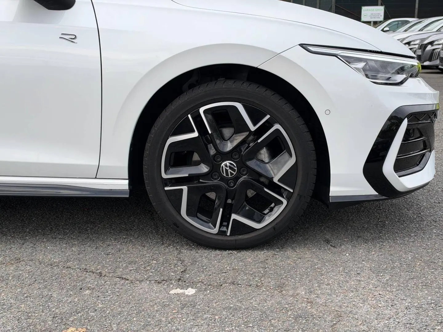 Gros plan sur la roue avant droite et partie avant blanche nacrée d'une Volkswagen Golf R-Line 2025 avec jante alu noire et argent.