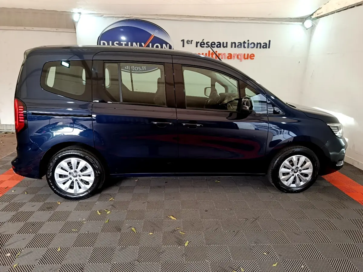 Profil côté droit d'un Renault Kangoo bleu 2023 garé en intérieur sur sol à damiers noirs et rouges.