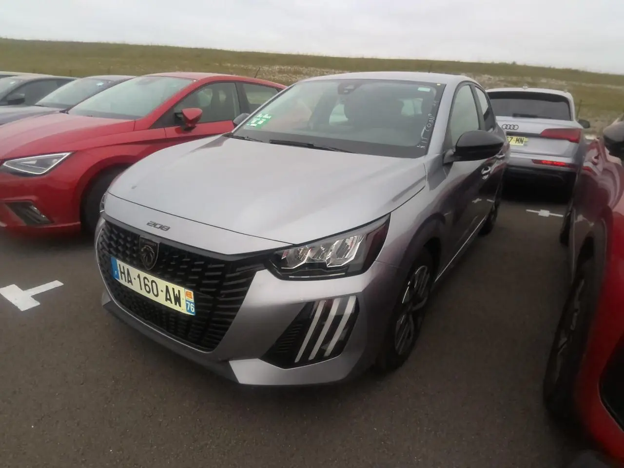 Peugeot 208 gris clair vue 3/4 avant avec calandre noire et feux à signature LED en forme de griffes.