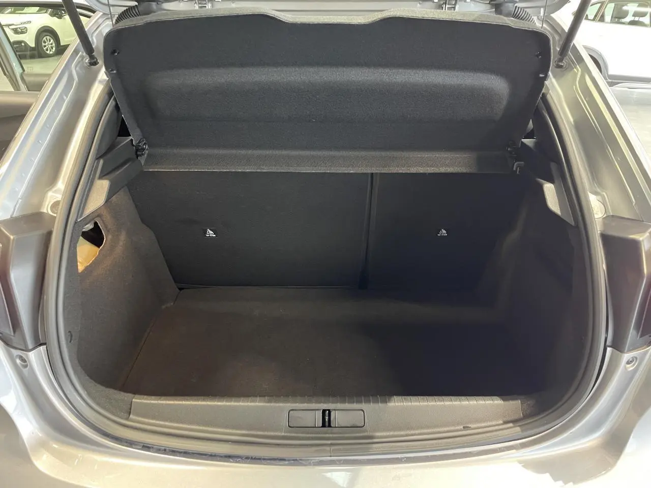 Coffre ouvert vu de l'arrière d'une Peugeot 208 gris clair, montrant l'espace de rangement vide et la trappe de compartiment.